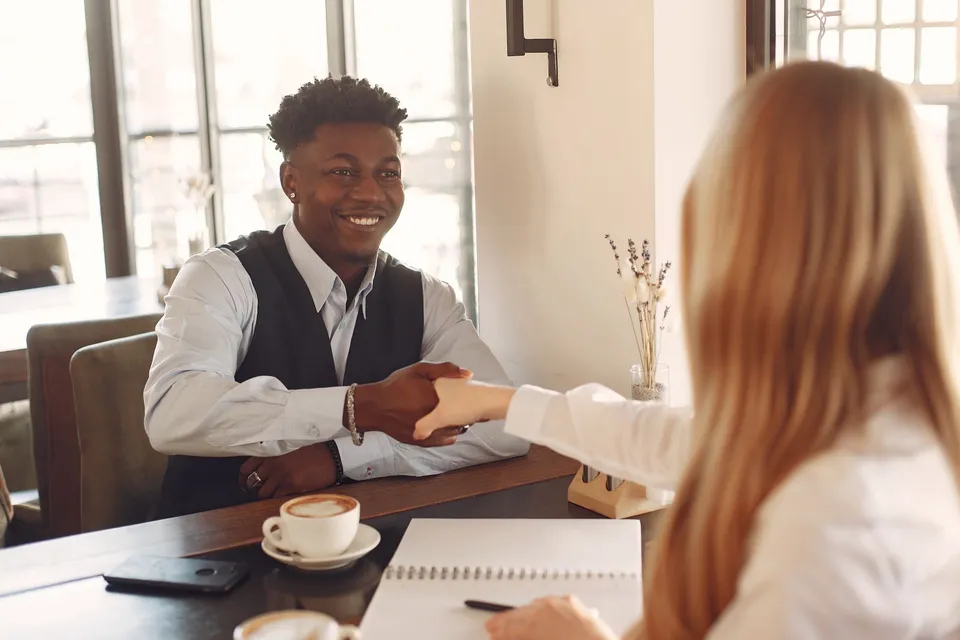 Two adults shaking hands across a table in a coffee shop, fostering a business connection.