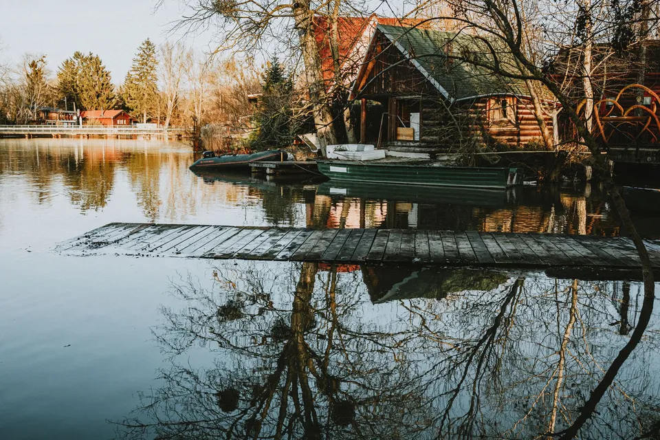 Rustic lakeside view featuring wooden cabins, boats, and reflections in tranquil waters.