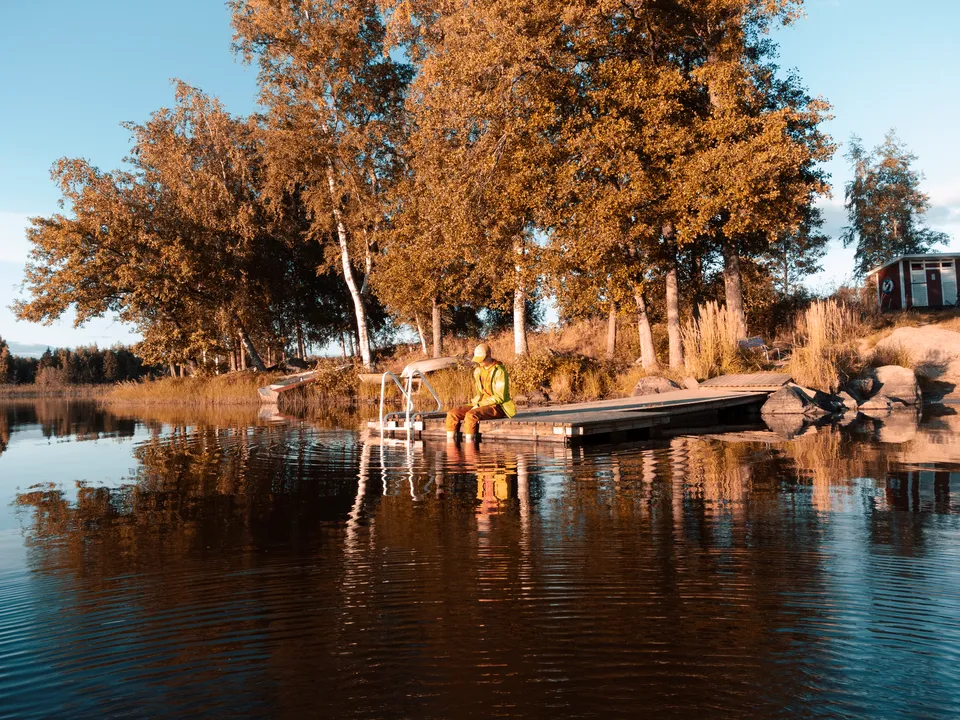 A person sits alone on a wooden dock by a lake surrounded by vibrant fall foliage.
