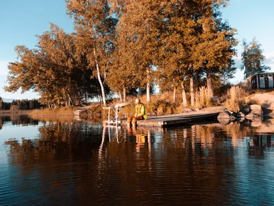 A person sits alone on a wooden dock by a lake surrounded by vibrant fall foliage.