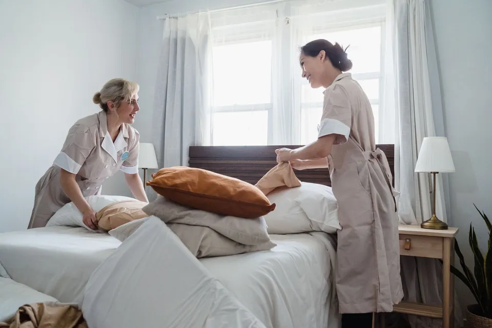 Two female housekeepers making a bed together in a well-lit room with tidy decor, showcasing teamwork.